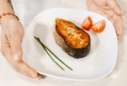 Close-up of grilled salmon served with tomatoes and chives on a plate, held by tattooed hands.